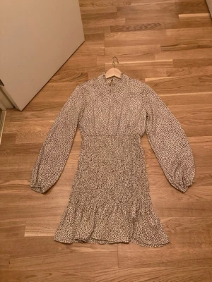 Prickig kortklänning med volang - Supersöt kortklänning med svart beige  prickigt mönster, långärmad och med volangkant nertill. Klänningen har hög hals och smockad midja som ger en snygg siluett. Perfekt för dig som gillar trendiga detaljer och en feminin look. Ny!!!