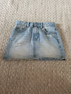 Ljusblå jeanskjol från Stradivarius - Trendig ljusblå jeanskjol från Stradivarius med klassisk femficksdesign och slitna detaljer framtill. Kjolen har bälteshällor och knappgylf, perfekt för en avslappnad och cool stil. Passar till sneakers eller boots för en casual look.