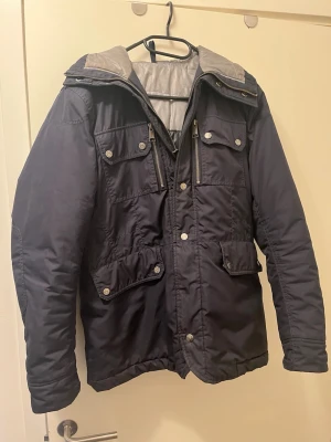 Mörkblå parkas från j Lindeberg - Snygg mörkblå parkas från J.Lindeberg  med flera fickor framtill, dragkedja och tryckknappar. Materialet är slitstarkt och vattenavvisande, perfekt för kalla dagar. Stilsäker design med detaljerade logoknappar.