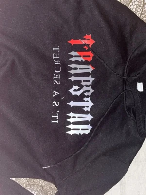 Svart hoodie med tryck 'Trapstar' XL - Svart hoodie med huva och snörning, stor tryckt text 'Trapstar' sopm går i vitt, grått och rött på bröstet. Under står det 'IT'S A SECRET' i vit och röd text. Perfekt för dig som gillar streetwear och vill sticka ut och den är i asiatisk storlek så den passar nog dem som har L eller M också XL