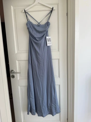 Blå långklänning med öppen rygg och knytning  - Supersnygg blå långklänning i satin med smala axelband som korsas i ryggen. Klänningen har öppen rygg och en knytdetalj baktill. Klänningen är aldrig använd med prislapp kvar. Perfekt till bal eller bröllop eller annan festlighet.