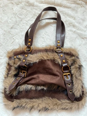 Brun handväska med fuskpäls och spännen - Cool brun handväska med detaljer i fuskpäls och två justerbara axelband i mörkbrunt konstläder. Väskan har dekorativa spännen och nitar i guldtonad metall. Perfekt för dig som gillar en unik och trendig look.