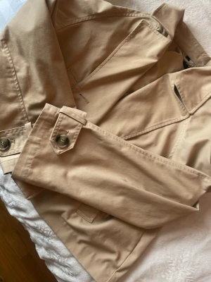 Beige trenchcoat med klassiska detaljer - Stilren beige trenchcoat med knappar och axelklaffar. Jackan har snygga detaljer vid ärmslut och fickor, samt klassisk krage. Perfekt för dig som vill ha en tidlös och trendig look. Materialet är slätt och känns bekvämt.