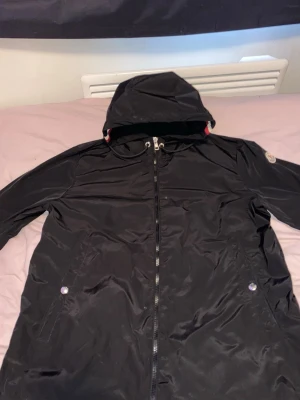 Svart Moncler vindjacka med huva - Snygg svart vindjacka från Moncler med huva och dragkedja framtill. Jackan har två fickor med tryckknappar och en randig detalj i blått, vitt och rött på insidan av huvan. Perfekt för blåsiga dagar och enkel att matcha med olika outfits.