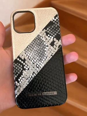 Mobilskal iPhone 15 iDeal of Sweden - Stilrent mobilskal med svart, silver och ormmönstrad design från iDeal of Sweden. Skalet har en lyxig känsla med struktur i materialet och passar perfekt för dig som vill ha något unikt till din mobil. Tillverkat i konstläder med metallogo. Nästan nytt, köpte fel modell, låda kommer till.