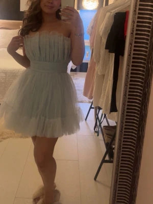 Ljusblå axelbandslös tyllklänning - Supersöt ljusblå kortklänning i tyll med volangdetaljer och bred midjeresår. Axelbandslös modell som ger en elegant och luftig känsla. Perfekt för dig som vill sticka ut med en romantisk och trendig look.