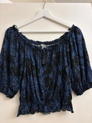 Blå offshoulder blus med knyt - Trendig blå offshoulder blus med svart och vitt mönster. Blusen har korta puffärmar, resår vid axlar och midja samt en liten knytning framtill. Perfekt för en avslappnad men snygg look.