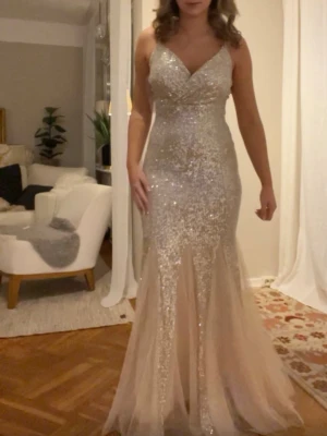 Glittrig långklänning i champagne - Säljer en magisk långklänning i champagnefärg med glittriga paljetter över hela klänningen. Klänningen har v-ringad omlottdesign upptill och markerad midja. Kjolen är utsvängd med transparenta partier nedtill för extra wow-effekt. Perfekt för bal eller festkväll! Priset går att diskutera!!!!💞