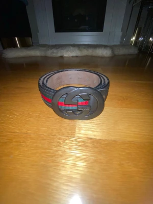 Svart Gucci bälte med GG-spänne - Snyggt svart bälte från Gucci med det klassiska GG-spännet i svart metall. Bältet har en randig detalj i rött och grönt längs mitten, vilket ger en ikonisk look. Tillverkat i skinn och passar perfekt till jeans eller byxor.