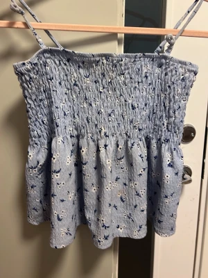 Blommig ljusblå linnetopp - Supersöt ljusblå linnetopp med smala axelband och blommigt mönster i vitt och blått. Toppen har smock upptill och ett flowy fall nedtill, perfekt för varma dagar. Materialet känns lätt och luftigt.