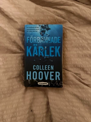 Förbannade kärlek av Colleen Hoover - En roman med blått omslag och stora turkosa bokstäver, skriven av Colleen Hoover. Boken heter 'Förbannade kärlek' och har ett omslag med vattenbubblor som ger en dramatisk känsla. Perfekt för dig som gillar romantik och starka känslor.