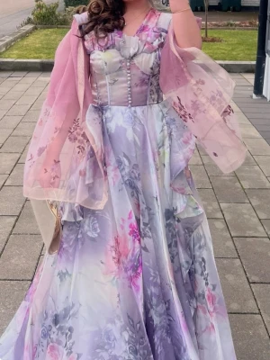 Bal klänning - Lång blommig klänning med volanger - Romantisk långklänning i pastellfärger med blommigt mönster och volangdetaljer. Klänningen har v-ringning, knappar framtill och är ärmlös. Materialet känns som satin eller polyester och den har en elegant, flytande siluett. Perfekt för sommarens speciella tillfällen.