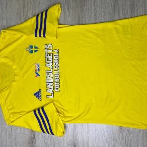 Gul Landslagets fotbollströja Adidas - Gul fotbollströja från Adidas med blå detaljer och tryck 'LANDSLAGETS FOTBOLLSSKOLA' på bröstet. Svenska fotbollsförbundets märke och Intersport-logga framtill. Klassisk rund hals och korta ärmar med blå ränder.