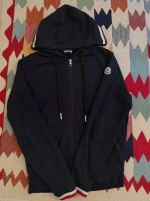 Svart hoodie från Moncler med dragkedja - Stilren svart hoodie från Moncler med hel dragkedja, huva och snören. Detaljer i rött, vitt och blått på huva och ärmslut samt Moncler-logga på vänster ärm. Perfekt för en avslappnad och trendig look. ❗️Till er som är intresserade priset är fast kommer inte gå ner i pris❗️