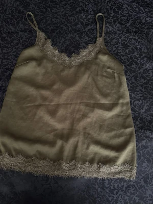 Olivgrönt linne med spetsdetaljer M - Snyggt olivgrönt linne med tunna axelband och spetsdetaljer längs både ringning och nederkant. Perfekt för mysiga kvällar hemma. Materialet känns mjukt och lätt mot huden, och spetsen ger en extra touch av stil.