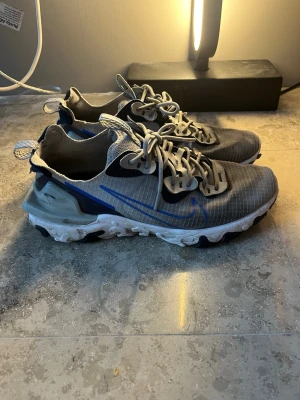Nike Reacts grå/blå sneakers - Nike React sneakers i grått med blå detaljer och vit sula. Skorna har genomskinlig mesh-överdel, reflekterande detaljer. Snygg design med synliga sömmar och unika snörhål, smutsiga men kan rengöra med skoprodukter innan säljning. Pris kan diskuteras vid snabb affär