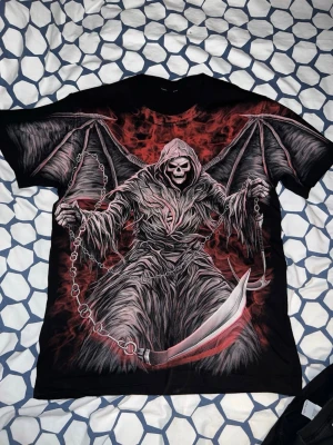 Svart t-shirt med Grim Reaper tryck - Säljer en svart t-shirt med ett stort, detaljerat tryck av Grim Reaper med vingar och lie i röd och grå färg. T-shirten har rund hals och korta ärmar. Perfekt för dig som gillar mörkare motiv och vill sticka ut.