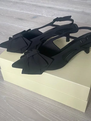 Svarta pumps med rosett och öppen häl - Stilrena svarta pumps med spetsig tå, stor dekorativ rosett framtill och öppen häl med justerbart spänne. Skorna har en låg klack och är tillverkade i tygmaterial. Helt oanvända endast testade, storlek 37. Nypris 399kr