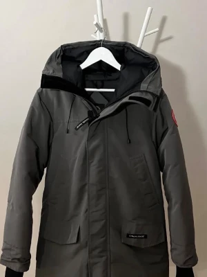 Grå parkas från Canada Goose - Snygg grå parkas från Canada Goose med klassisk röd logotyp på ärmen. Jackan har huva med dragsko, stora fickor framtill och dragkedja med vindslå. Perfekt för kalla dagar och riktigt varm tack vare det tåliga materialet.