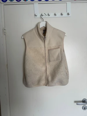 Beige fleeceväst med dragkedja - Mysig beige fleeceväst med hög krage och dragkedja framtill. Västen har en bröstficka på vänster sida och är tillverkad i ett mjukt, teddy-liknande material. Perfekt för lager-på-lager och enkel att matcha med olika outfits.