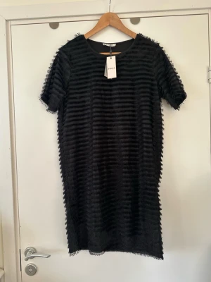Svart klänning med fransar, helt NY - Superfin helt NY klänning från Pieces i storlek M. Unikt texturerat mönster tyg med horisontella ränder av fransar. Klassiska korta ärmar och rund halsringning. Trendig rak passform som faller löst över kroppen och slutar ovanför knäna. Fodrad. Verkligen en pang-klänning, men tyvärr för liten för mig. Helt NY med etikett.   Skickas mot porto, kan även hämtas i Stockholm. 