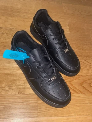 Nike Air Force 1 Low helsvarta - Snygga helsvarta Nike Air Force 1 Low sneakers i läder med klassisk siluett och perforerad tåbox. Skorna har svarta skosnören, metall-detalj med 'AF1' och diskret Nike-logga på sidan. Perfekta för dig som gillar stilrena och tidlösa sneakers.