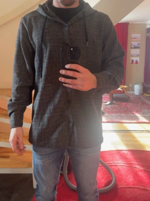 Mörkgrå overshirt med huva - Snygg mörkgrå overshirt med diskret rutmönster och huva med dragsko. Knäppning framtill och avslappnad passform. Perfekt lager på lager-plagg för en casual look. Tillverkad i mjukt tyg som känns bekvämt hela dagen. Sitter står XXL men den sitter som M/L