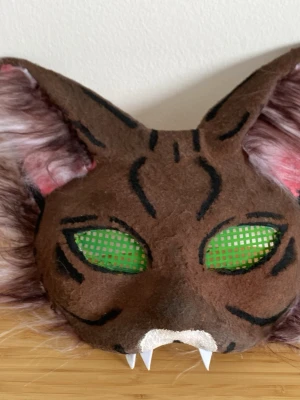 Handgjord kattmask med gröna ögon - Unik kattmask för cosplay eller maskerad, tillverkad i brunt tyg med svarta ränder och stora spetsiga öron. Masken har gröna nätögon, vita huggtänder och rosa detaljer i öronen. Fästs med elastiskt band på baksidan. Perfekt för dig som vill sticka ut!