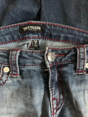 True Religion Joey Flare Jeans - Säljer ett par mörkblå True Religion Joey Ultra Low Rise Flare jeans med röda kontrastsömmar och stora bakfickor med signaturbrodyr. Perfekta för dig som gillar statement-jeans! Jag säljer dessa då de tyvärr är för små för mig. Byxorna är bara använda 2-5 gånger men är slitna längst ner pga att jag är kort. 