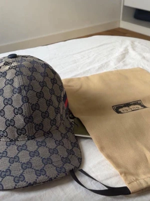 Gucci beige GG-mönstrad keps - Säljer en beige keps från Gucci med klassiskt GG-monogram i mörkblått över hela kepsen. Kepsen har böjd skärm och justerbar rem bak. På sidan finns en röd och marinblå rand. Materialet är canvas och kepsen kommer med original dustbag.