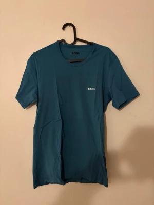 Blå kortärmad t-shirt från BOSS - Snygg blå t-shirt från BOSS med enkel och stilren design. T-shirten har rund halsringning och korta ärmar. Liten vit BOSS-logga på bröstet. Perfekt basplagg i mjuk bomull som passar till det mesta.