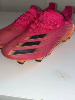 Adidas rosa fotbollsskor med dobbar - Säljer ett par snygga fotbollsskor från Adidas i en stark rosa färg med svarta och guldiga detaljer på sidorna. Skorna har klassiska dobbar för bra grepp på planen och är tillverkade i ett lätt syntetmaterial. Perfekta för dig som vill sticka ut på fotbollsplanen!
