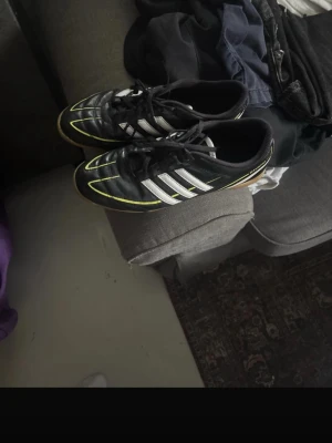 Adidas svartvita fotbollsskor - Säljer ett par klassiska fotbollsskor från Adidas i svart med vita ränder och gula detaljer. Skorna har snörning och låg profil, perfekta för spel på gräsplan. Materialet är syntet med textilfoder för bra komfort och passform.