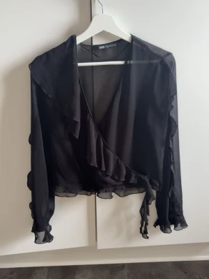 Svart omlottblus med volanger Zara - Snygg svart omlottblus från Zara i transparent, lätt material. Blusen har långa ärmar och volangdetaljer längs kanterna och ärmsluten. Perfekt för dig som gillar en trendig och feminin look med lite extra detaljer.