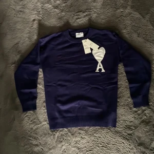 Ami Paris tröja - Snygg mörkblå stickad tröja från Ami Paris med deras ikoniska vita hjärta och A-logga på bröstet. Tröjan har rund hals, ribbade muddar och är långärmad. Perfekt för dig som vill ha en stilren och trendig look.