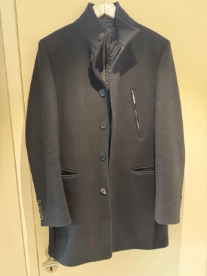 Svart rock från Dressmann, storlek 46 - Klassisk svart rock från Dressmann med ståkrage och knappstängning. Rocken har tre framfickor, varav en bröstficka med dragkedja, samt dekorativa knappar vid ärmsluten. Tillverkad i ett kraftigare tyg med slät finish och helfodrad insida.