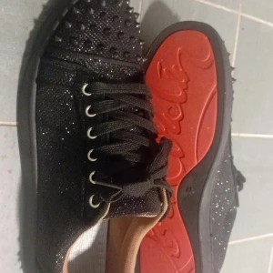 Svarta glittriga sneakers med nitar - Säljer ett par svarta sneakers från Christian Louboutin med glittrigt tyg och svarta nitar på tån och hälen. Skorna har snörning och den klassiska röda sulan med logotyp. Perfekt för dig som vill sticka ut med en edgy och lyxig stil.