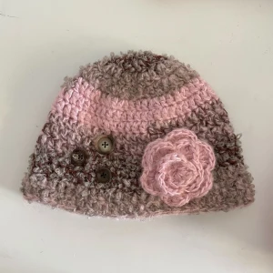 Handvirkad mössa med ros - Söt virkad mössa i rosa och bruna färger. Virkad med fluffigt garn, perfekt för det kalla vädret!❄️ Mått: längd: 19 cm, bredd (längst ner på mössan) : 27cm 💌 Skriv om ni har fler frågor!