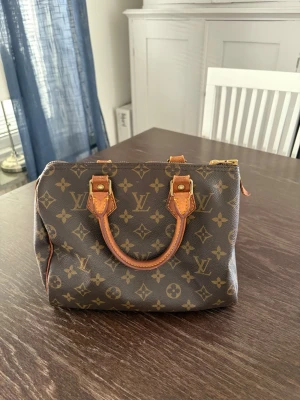 Louis Vuitton Speedy Monogram handväska - Klassisk Louis Vuitton Speedy handväska i brunt monogram canvas med detaljer i läder och guldfärgade metalldelar. Väskan har två korta handtag och dragkedja upptill. Ikoniskt LV-mönster över hela väskan och snygga kontrastsömmar. Speedy 25! 