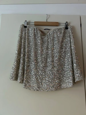 Glittrig silvertopp med paljetter - Topp i silver med glittriga paljetter över hela tyget. Toppen har en lätt utställd form och resår vid bysten för bekväm passform. Perfekt för dig som vill ha något som verkligen syns och reflekterar ljuset snyggt.