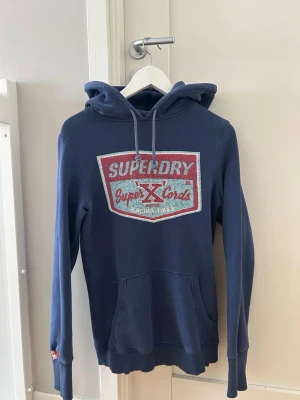 Blå hoodie från Superdry - Snygg blå hoodie från Superdry med stor logga och text på bröstet i rött och vitt. Klassisk känguruficka framtill och justerbar huva med dragsko. Mjuk insida och ribbade muddar vid ärmar och nederkant. Perfekt för en avslappnad streetwear-look. Storleken är M men är liten i designen bara så skulle säga de är Small