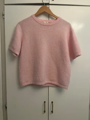Ljusrosa stickad t-shirt - Söt ljusrosa stickad t-shirt med korta ärmar och rund hals. Tröjan har en mjuk och fluffig struktur som ger en mysig känsla. Passar perfekt till jeans eller kjol för en avslappnad look.