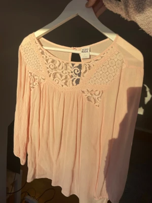 Ljusrosa blus med spets från Vero Moda - Söt ljusrosa blus från Vero Moda med vackra spetsdetaljer vid halsringningen och axlarna. Blusen har en lös passform och är tillverkad i ett tunt, luftigt tyg som ger en skön känsla. Perfekt för dig som gillar romantisk och feminin stil.