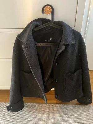 Svart ullkappa från 157 - Stilren svart ullkappa från 157 med klassisk krage, stora framfickor och knäppning framtill. Kortare modell som ger en modern look. Perfekt för kyliga dagar och enkel att matcha med olika outfits.
