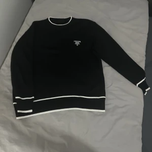 Svart tröja med vita detaljer från Prada - Snygg svart tröja från Prada med vita kontrastsömmar vid hals, ärmslut och nederkant. Rund halsringning och diskret logga på bröstet. Perfekt för en stilren och modern look.