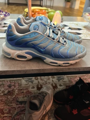 Nike Air Max Plus blå sneakers - Nike Air Max Plus sneakers i blått med vita och ljusblå detaljer. Skorna har den ikoniska vågiga designen på ovandelen, meshmaterial och synliga Air-enheter i sulan. Swoosh-logga på sidan och vita skosnören. Perfekta för dig som gillar streetwear och sportig stil.