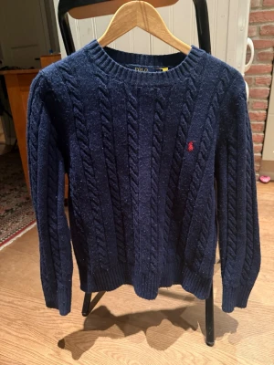 Mörkblå kabelstickad tröja Polo Ralph Lauren - Kabelstickad mörkblå tröja från Polo Ralph Lauren med rund halsringning och ribbade muddar. Tröjan har det klassiska röda Polo-logot broderat på bröstet och är tillverkad i mjukt material. Perfekt för dig som gillar tidlös och stilren design.