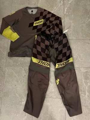 Thor motocross set brun/gul - Säljer ett motocross set från Thor i storlek L. Tröjan och byxorna är bruna med gul och svart rutigt mönster samt gula detaljer och logga. Tillverkad i slitstarkt syntetmaterial, perfekt för motocross eller enduro. Långärmad tröja och byxor med förstärkta knän. Det är i använt skick men funkar perfekt och inga större hål eller något som känns på banan. Nypris ligger runt 1299kr.