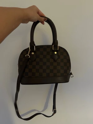 Louis Vuitton Väska - Ej äkta! Louis Vuitton väska i modellen Alma. Ett innerfack, ett hänglås och ett långt band tillkommer. Bra skick, men i sista bilden syns att ena spännet till det långa bandet lossnat, det går att fixa annars kan man ha den som på bilden eller ta bort bandet helt