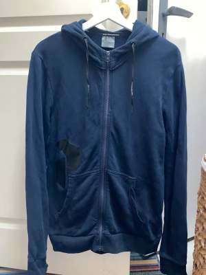 Mörkblå zip-hoodie från Peak Performance - Mörkblå hoodie med dragkedja från Peak Performance. Har huva med dragsko, två fickor fram och en svart detalj på vänster sida. Tillverkad i mjukt bomullsmaterial, perfekt för chill dagar. Klassisk och enkel design som funkar till det mesta. Storlek S